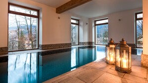 Une grande piscine intérieure offrant une vue dégagée sur les montagnes grâce à de larges baies vitrées, ainsi que deux lanternes illuminées placées au bord du bassin.
