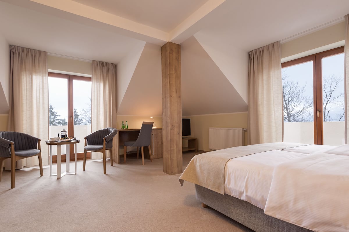 A hotel room with a large bed, two armchairs, a small table with a lamp, a TV, and a window with curtains.