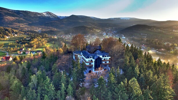 Une grande maison moderne, entourée d’une forêt dense, avec vue sur un petit village et des montagnes en arrière-plan.