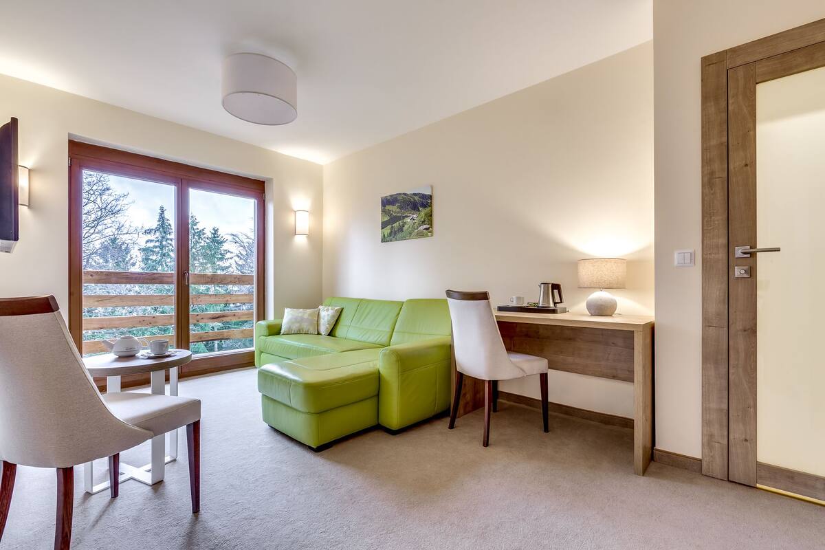 A modern living room with a green sofa, a white chair, a wooden desk, and a large window with a view of trees.