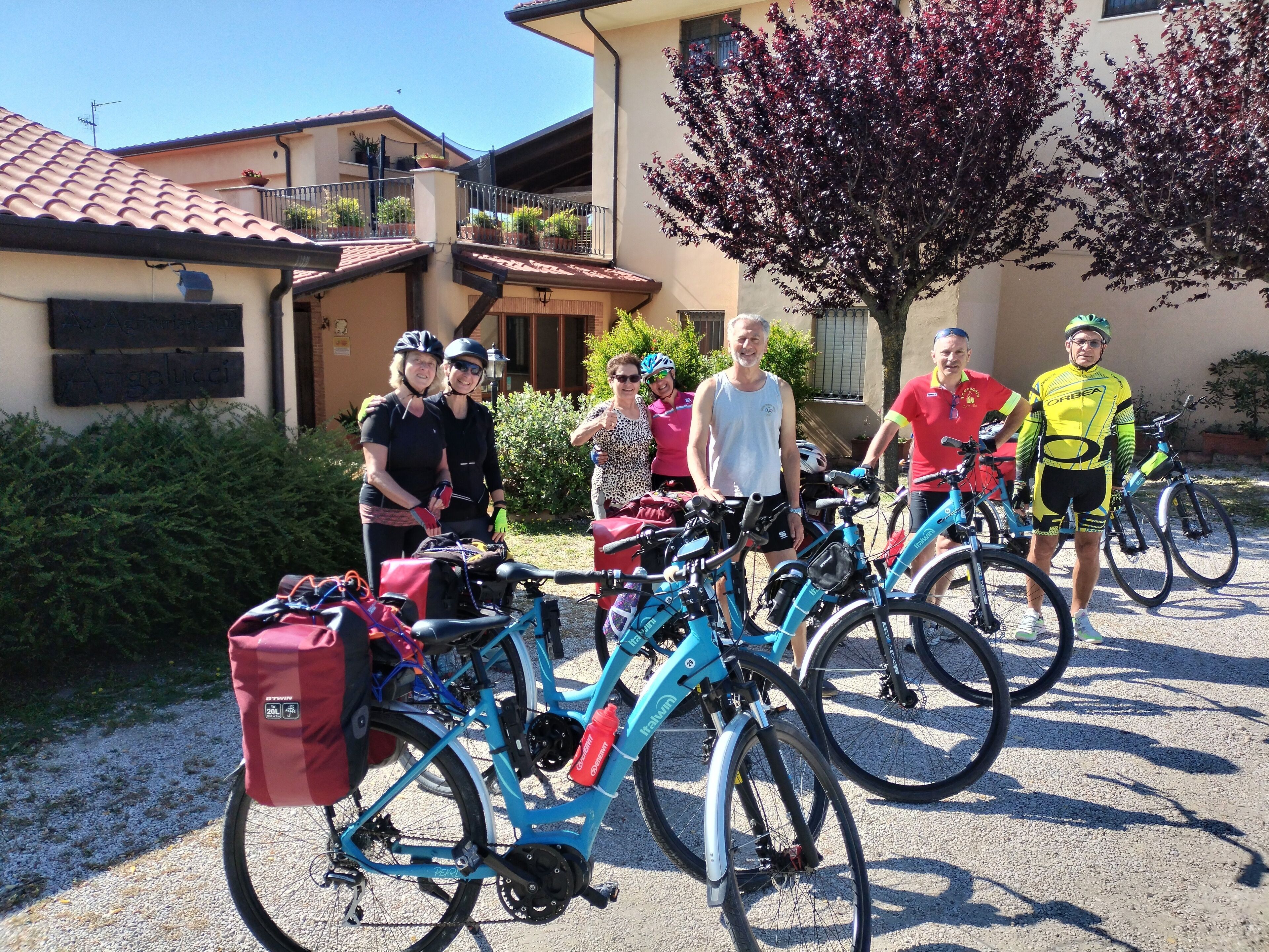 A group of cyclists with bicycles parked in front of a building with a tiled roof and a tree with red leaves.