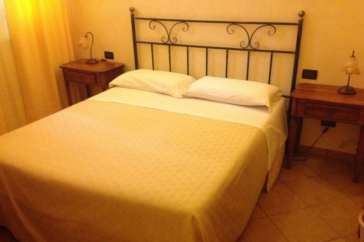 Una cama bien hecha con una colcha sencilla, dos mesitas de noche con lámparas y un cabecero simple.