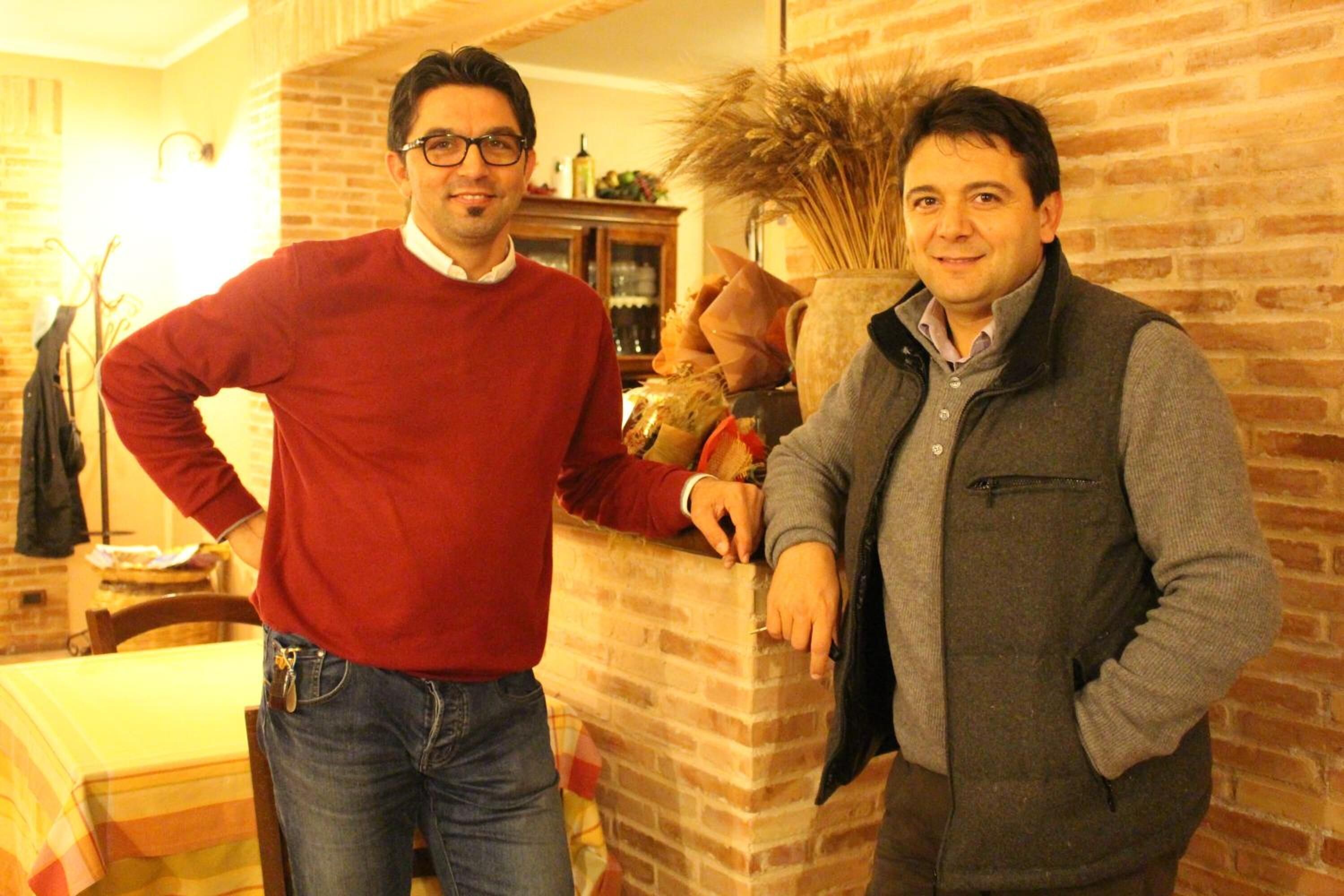 Dos hombres de pie en una habitación con paredes de ladrillo y una mesa con mantel a rayas.