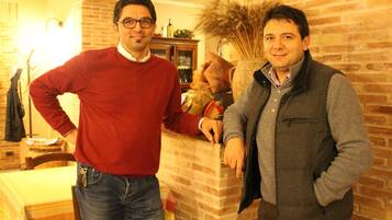 Dos hombres de pie en una habitación con paredes de ladrillo y una mesa con mantel a rayas.