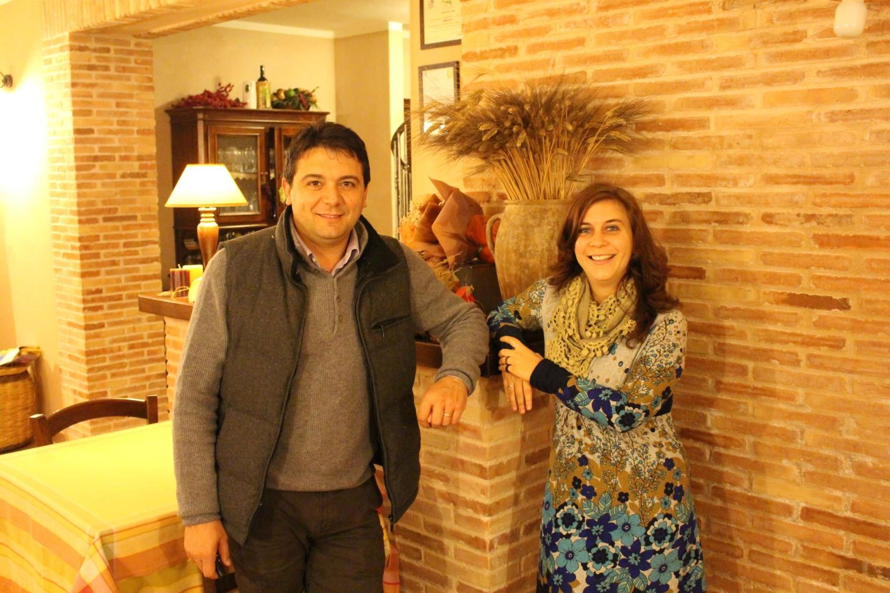 A man and woman standing in a room with brick walls, a wooden cabinet, and a table with a striped tablecloth.