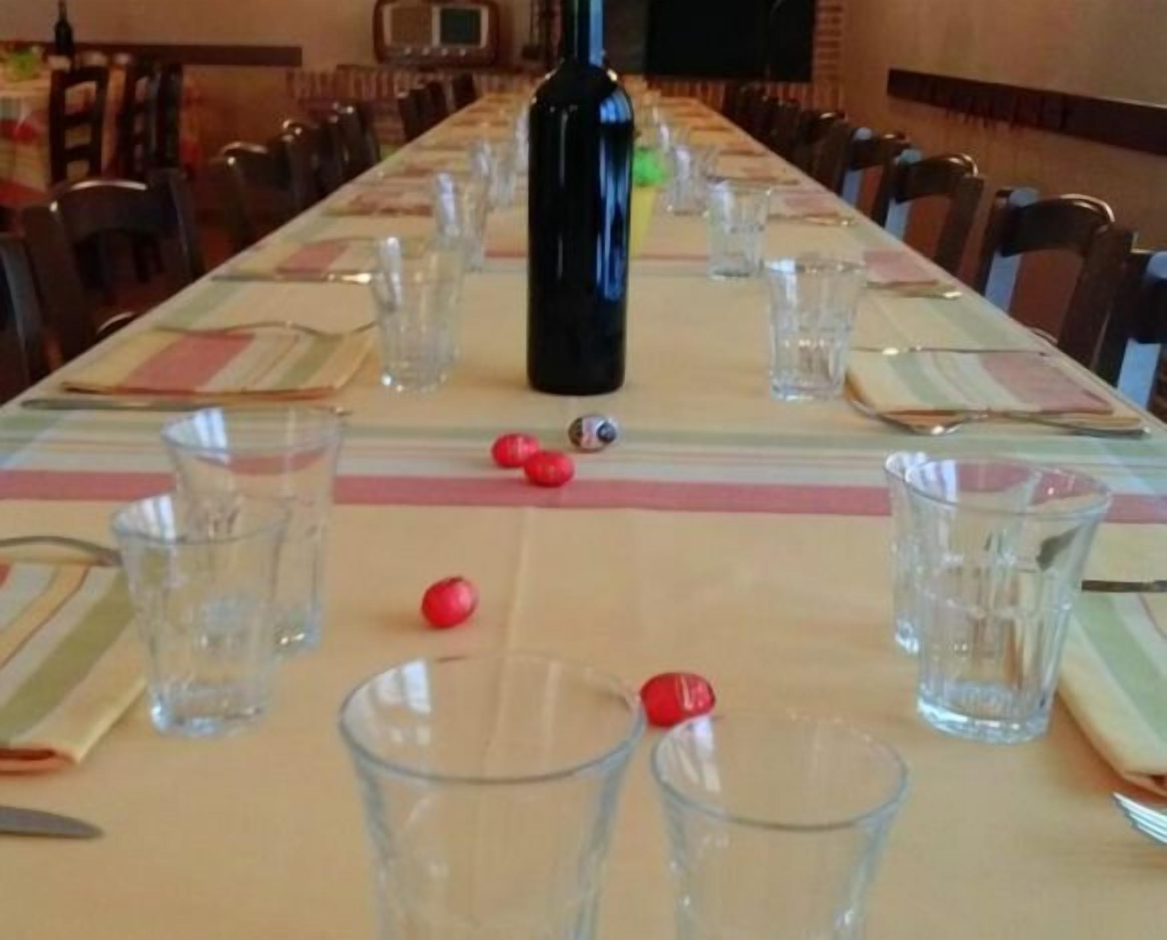 Una larga mesa de comedor dispuesta con vasos transparentes, platos y cubiertos, con una botella y algunos objetos rojos encima.