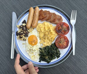 A plate of breakfast food including sausages, bacon, scrambled eggs, tomatoes, and greens.