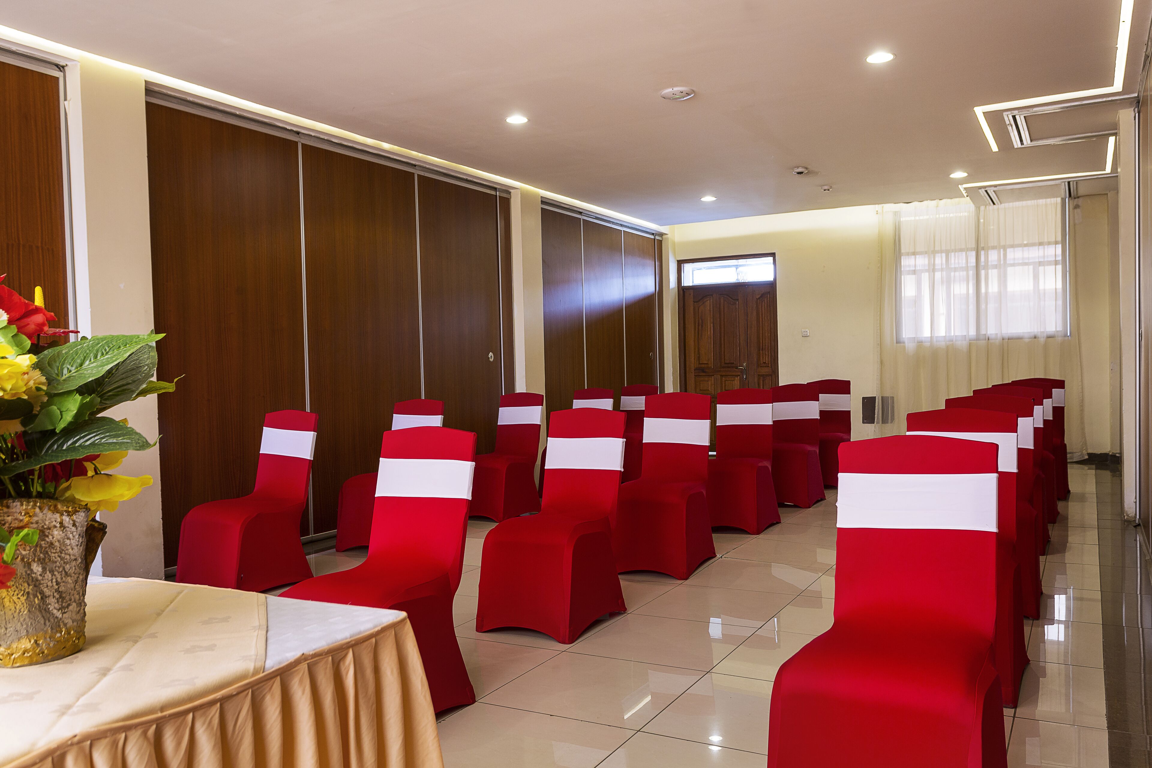 Uma sala de conferências com fileiras de cadeiras vermelhas, uma mesa com toalha bege e um vaso com flores amarelas.