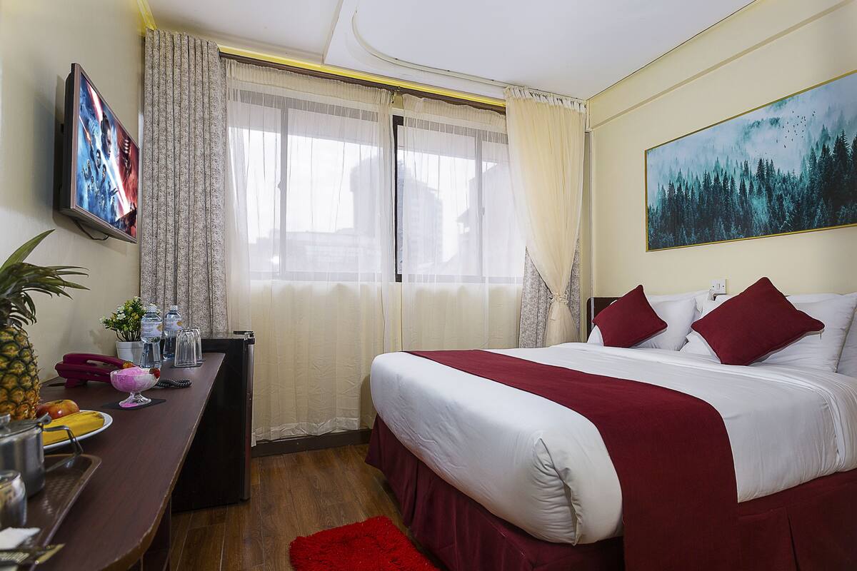 Habitación de hotel con una cama, un escritorio con computadora, una ventana con cortinas, un cuadro en la pared y una alfombra roja.