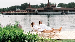 Ein Paar entspannt auf einer Holzterrasse an einem See, im Hintergrund ein traditionelles Gebäude.
