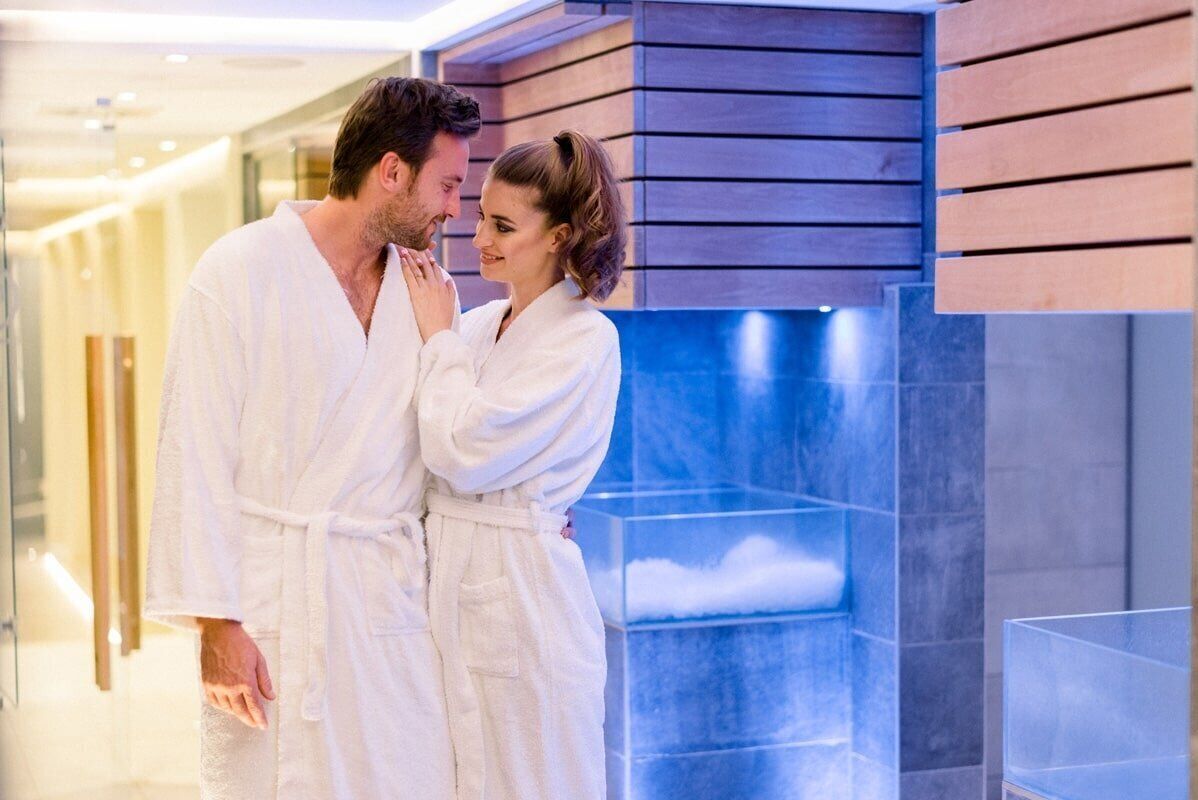 A couple in white bathrobes standing in a modern hotel corridor with wooden paneling and a glass enclosure.