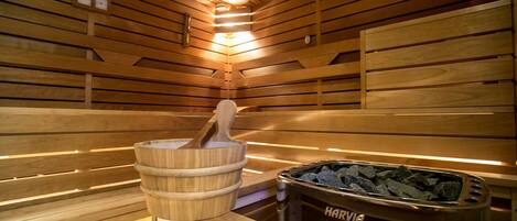A sauna with wooden benches, a wooden bucket with a mallet, and a container labeled "HARVIA" for stones.