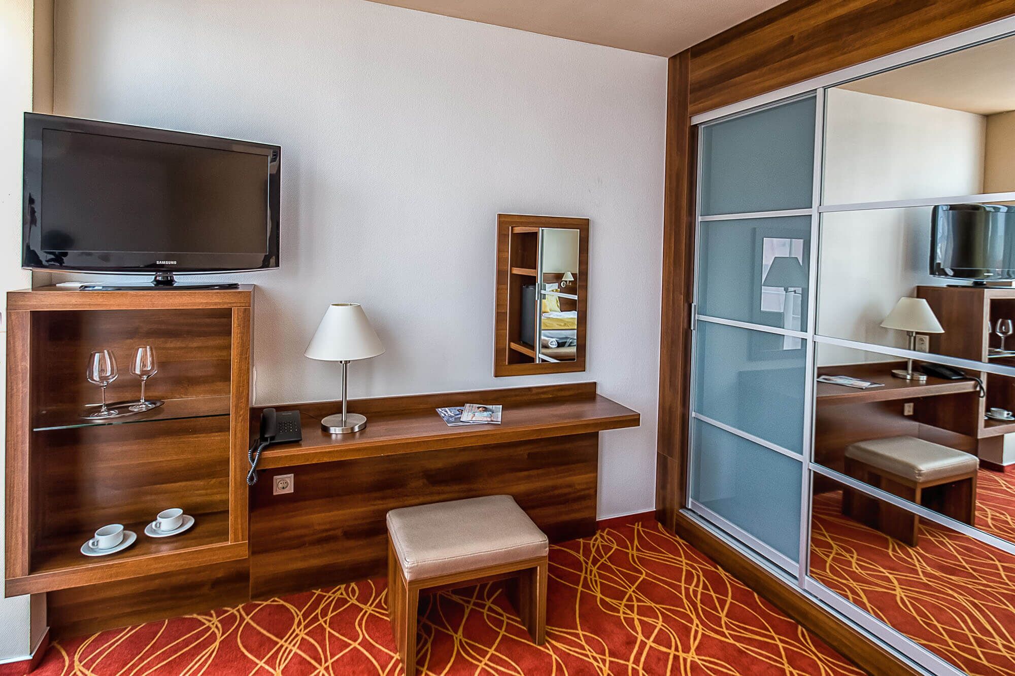 A hotel room with a TV, a desk with a lamp, a mirror, and a seating area with a red patterned carpet.