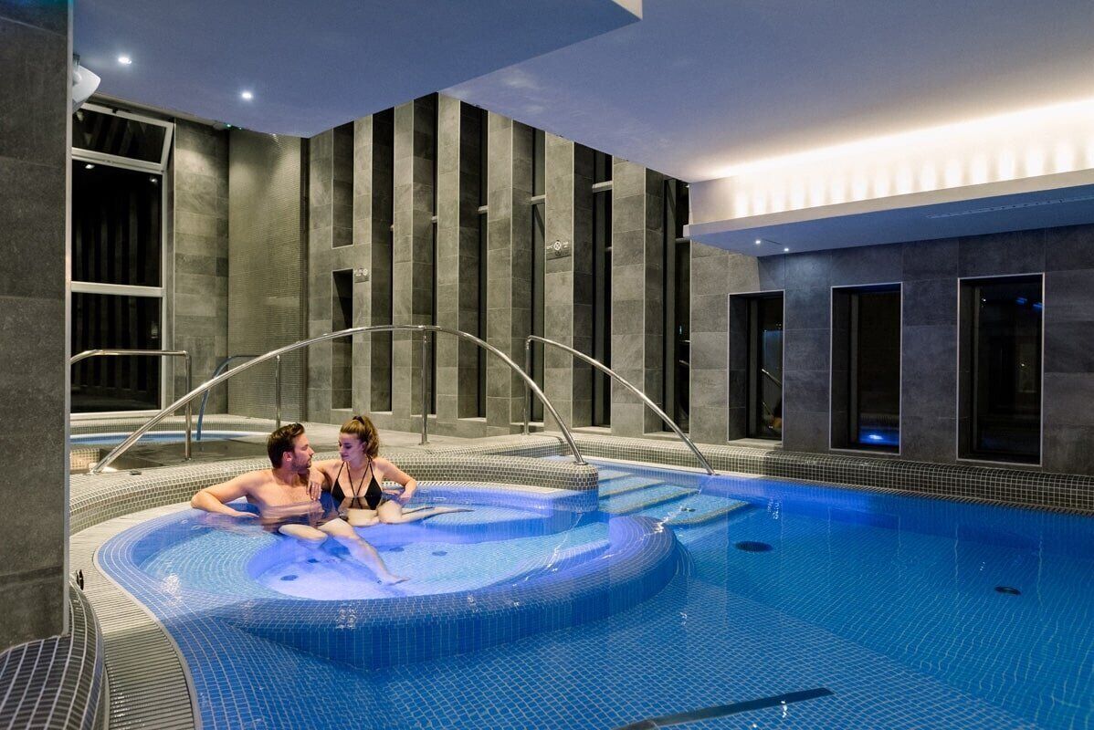 Una piscina interior moderna con zona de hidromasaje, rodeada de paredes de piedra oscura y grandes ventanales.