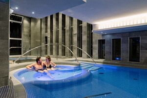 Una piscina interior moderna con zona de hidromasaje, rodeada de paredes de piedra oscura y grandes ventanales.