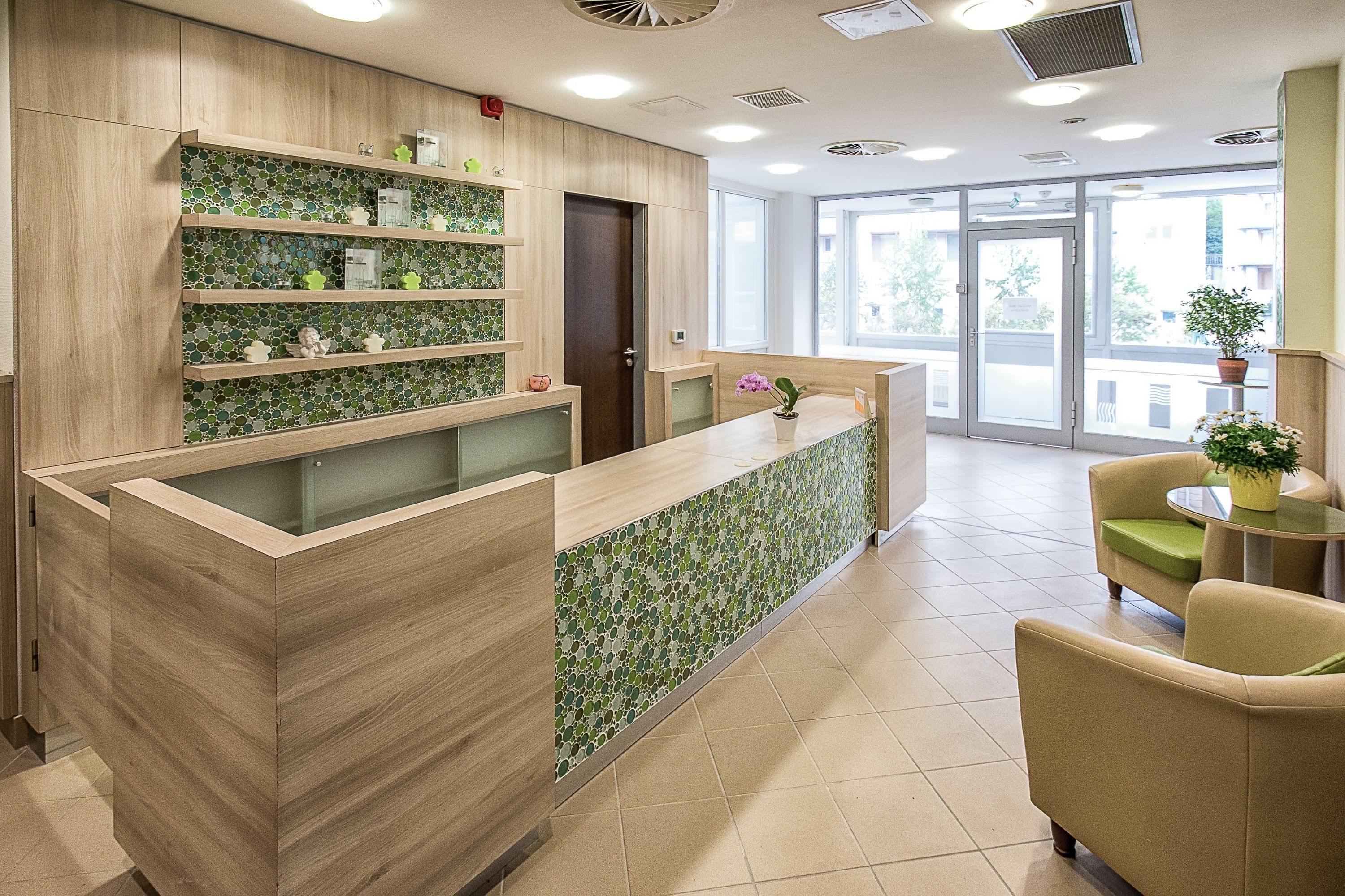 Zona de recepción con mostrador de madera, estanterías con azulejos verdes y vistas al exterior a través de amplios ventanales.