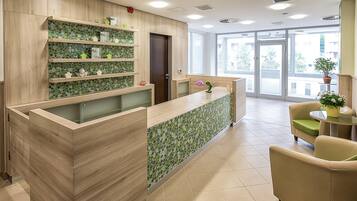 Zona de recepción con mostrador de madera, estanterías con azulejos verdes y vistas al exterior a través de amplios ventanales.