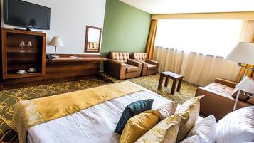 A hotel room with a bed, two armchairs, a small table, a TV, and a minibar.