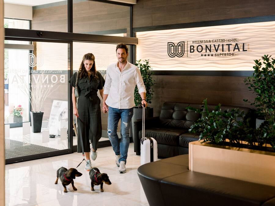 Un espace d’accueil avec une enseigne indiquant « BonVital Wellness & Gastro Hotel », un canapé en cuir noir, une plante en pot et un chien tenu en laisse.