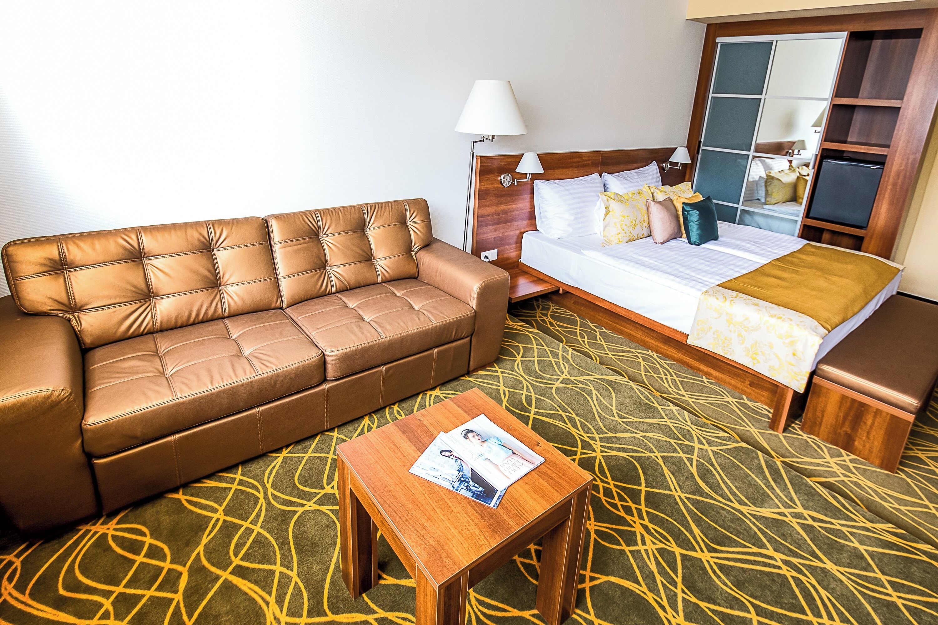 Une chambre d’hôtel avec un canapé en cuir, une table basse en bois sur laquelle sont posés des magazines, un lit recouvert d’une couvre-lit à motifs et une table de chevet avec une lampe.