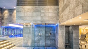 Ein moderner Aufzug mit Glastür und blauer Beleuchtung, umgeben von Steinwänden und einer Treppe.