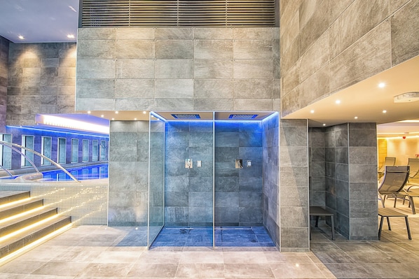 Un ascenseur moderne doté d’une cabine en verre et d’un éclairage bleu, entouré de murs en pierre et d’un escalier.