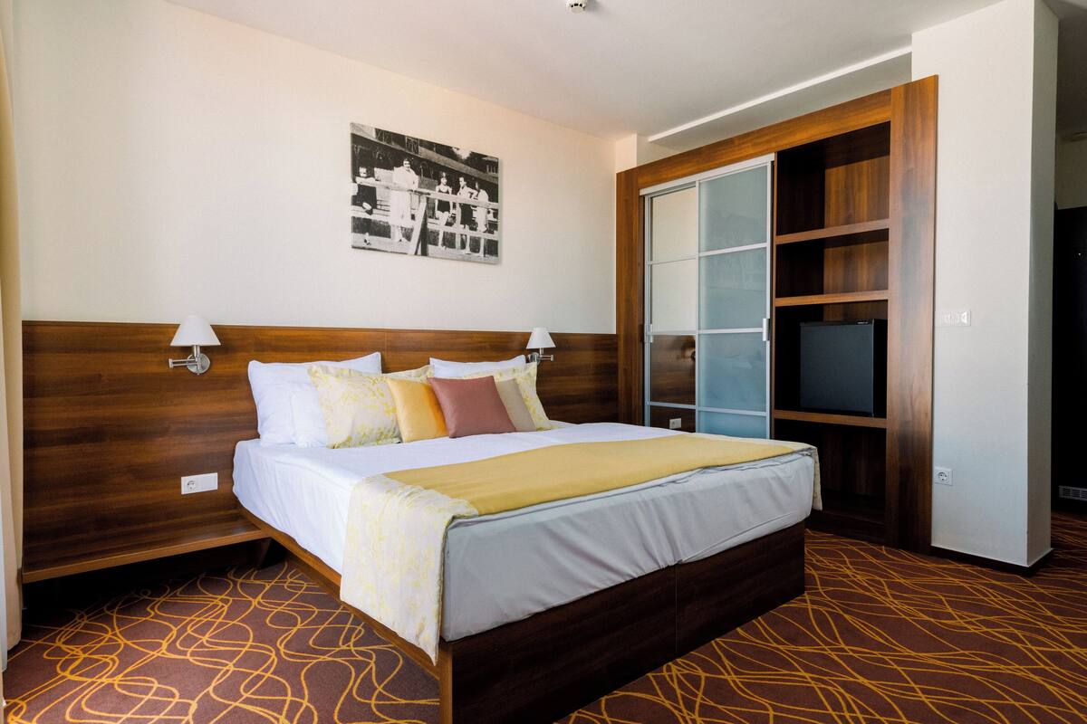 Ein Hotelzimmer mit hölzernem Kopfteil, einem Bett mit weißer Bettwäsche und mehreren Kissen, einem Nachttisch mit Lampe, einem Wandbild und einem Kleiderschrank mit Spiegel und Fernseher.