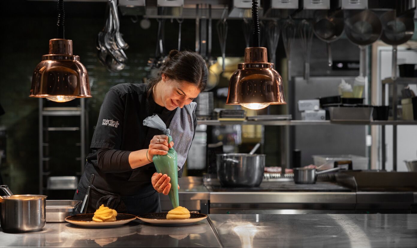 A person in a kitchen preparing food, with hanging lights and kitchen equipment in the background.
