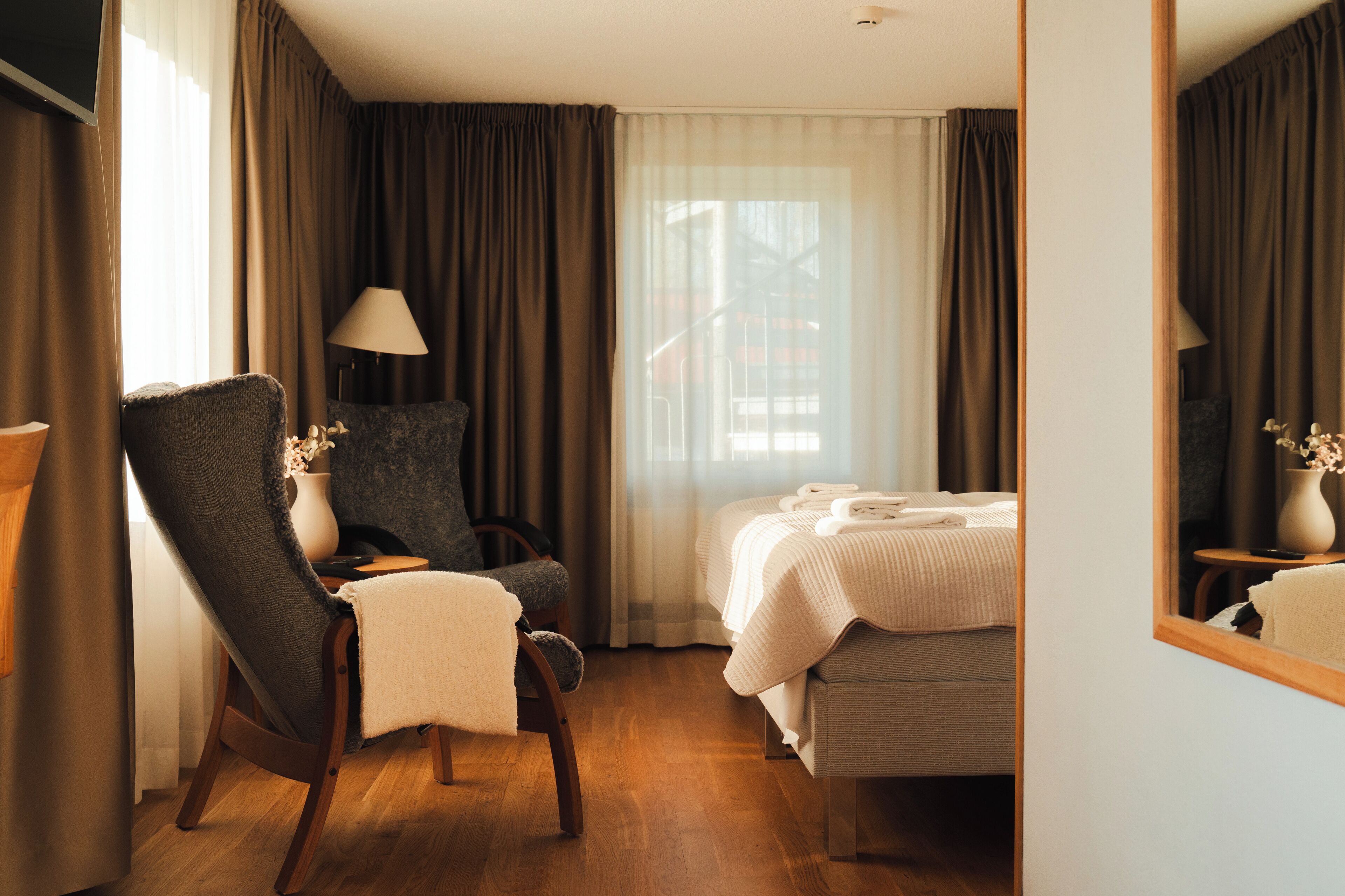 Ett hotellrum med en säng, en stol, en lampa och ett fönster med gardiner.