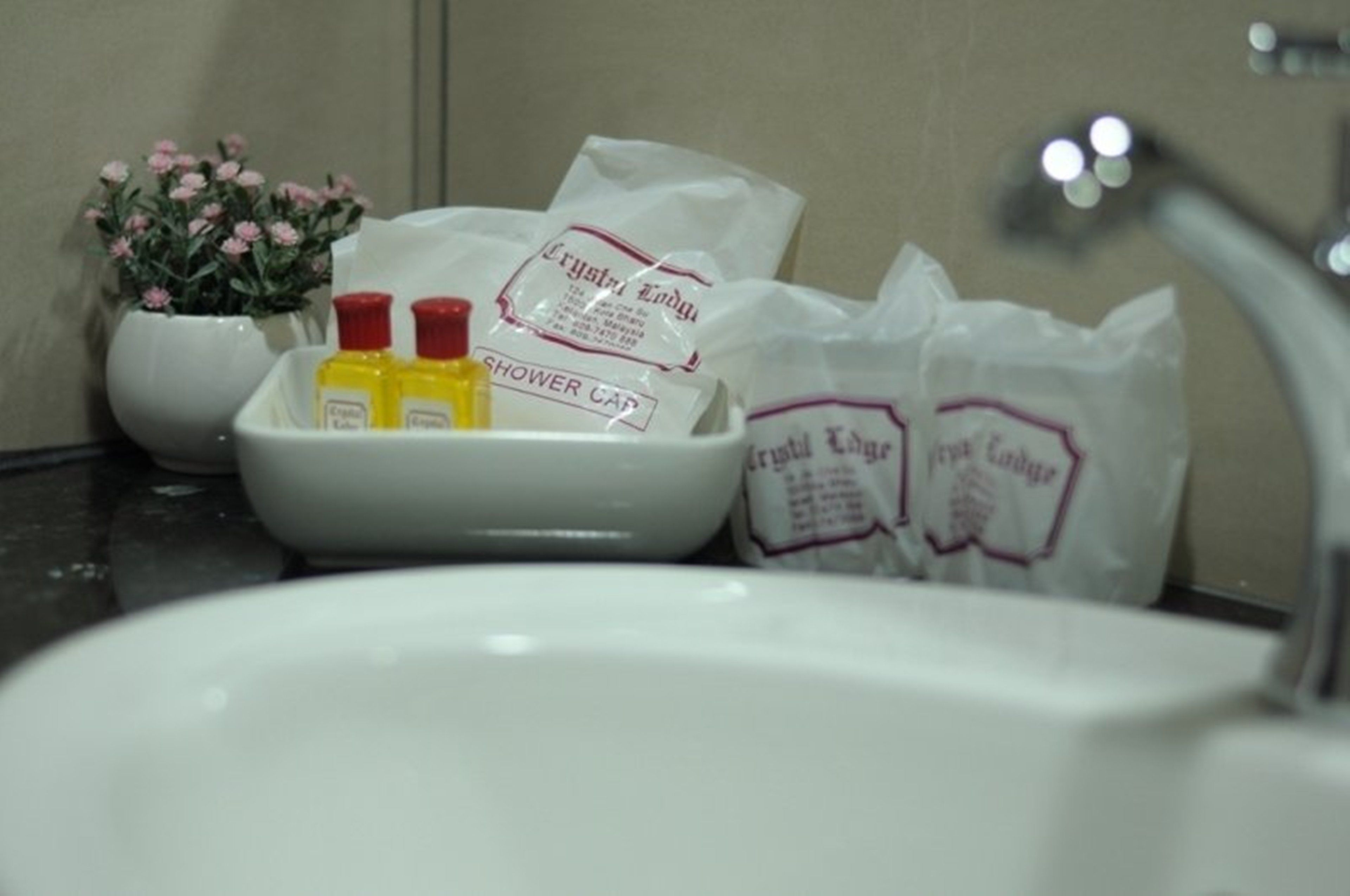 A bathroom sink with a white basin, a potted plant with pink flowers, and a tray with two yellow bottles and a white bag labeled "SHOWER CAP".