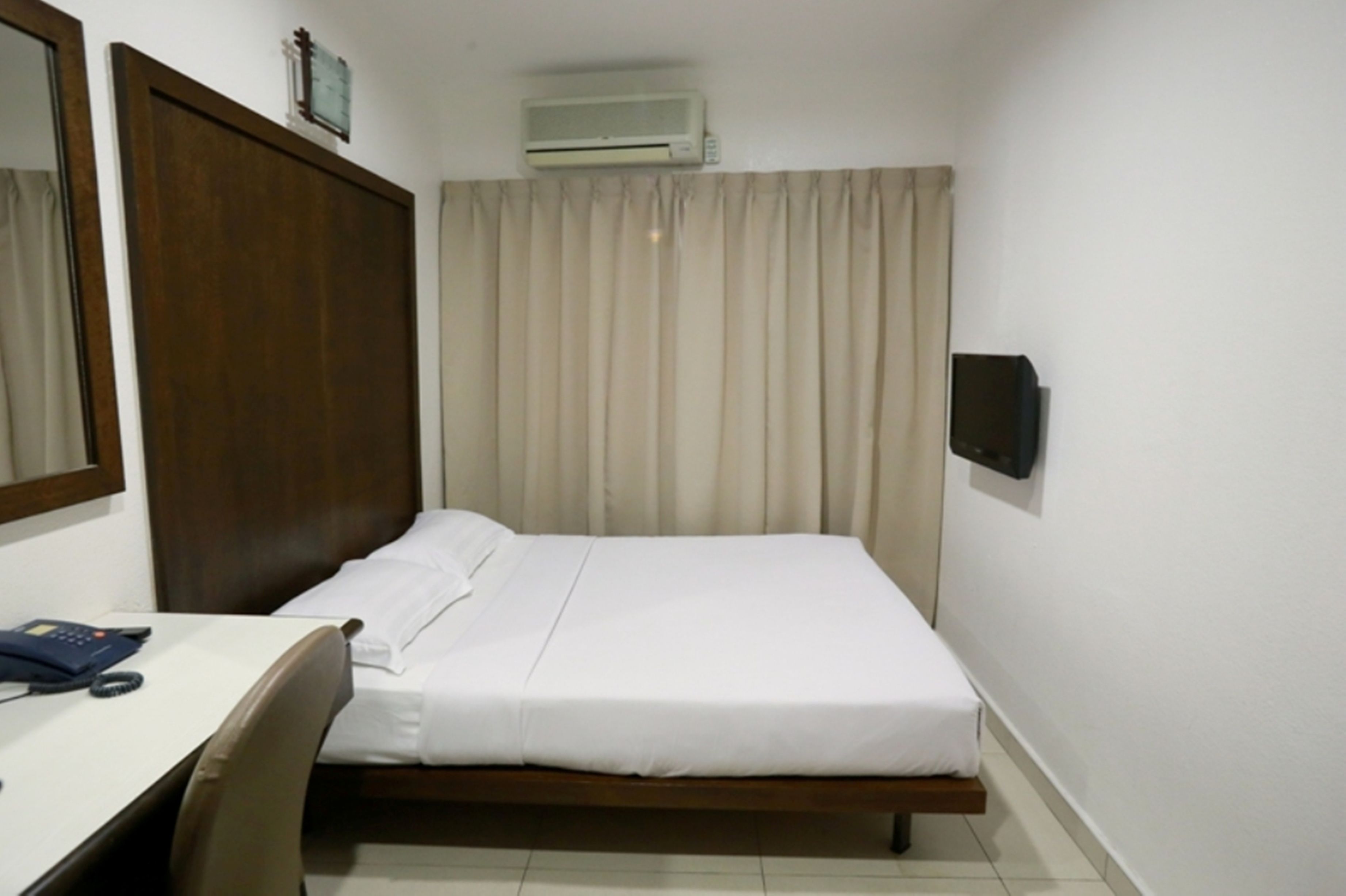 A hotel room with a bed, a desk with a telephone, a wall-mounted air conditioner, and a curtain.