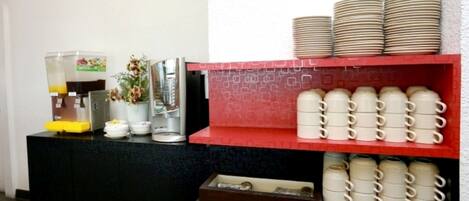 A buffet setup with a coffee station, a fruit dispenser, and neatly arranged plates and cups.