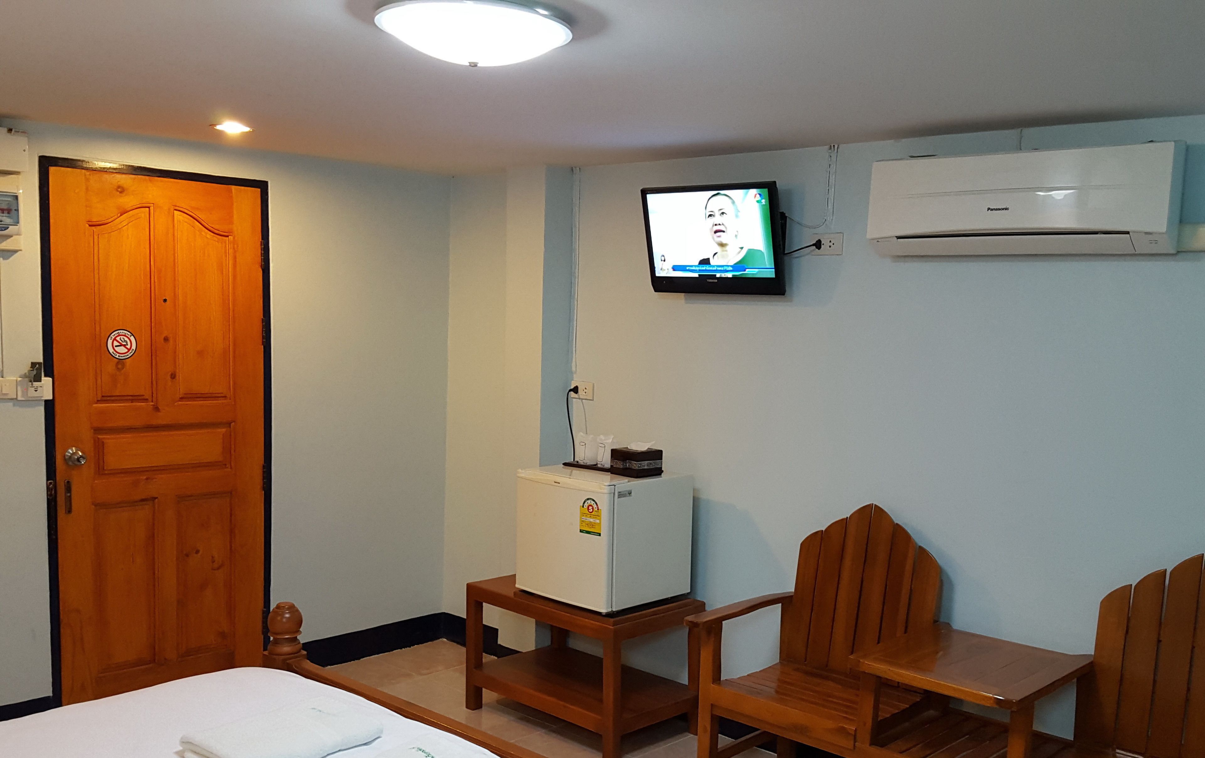 A hotel room with a wooden door, a small refrigerator, a television mounted on the wall, and a wooden chair.
