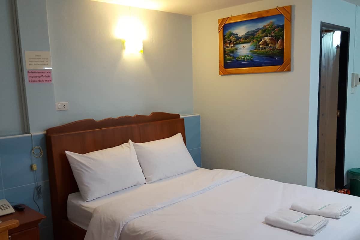 A neatly made bed with white linens and pillows, a wooden headboard, a painting of a scenic landscape on the wall, and a small table with a telephone and a charger.