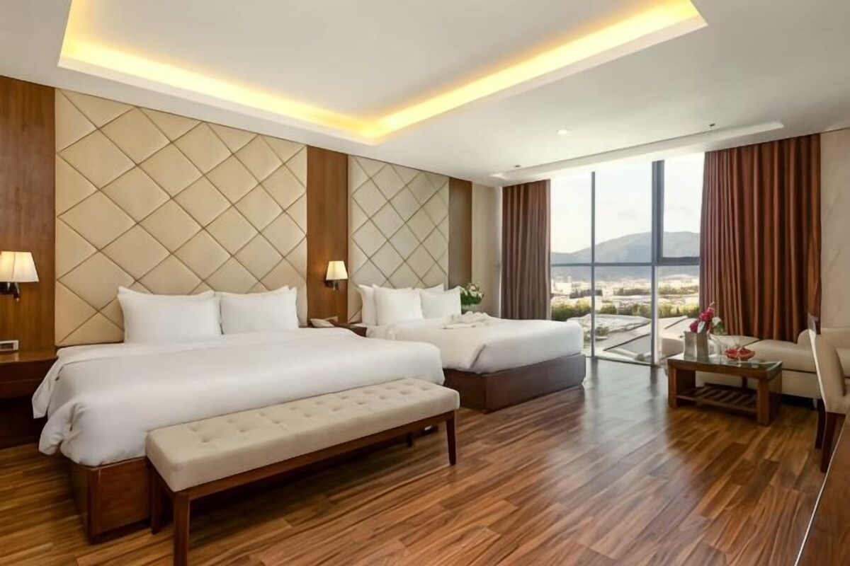 A hotel room with two beds, a bench, a sofa, and a view of the city.