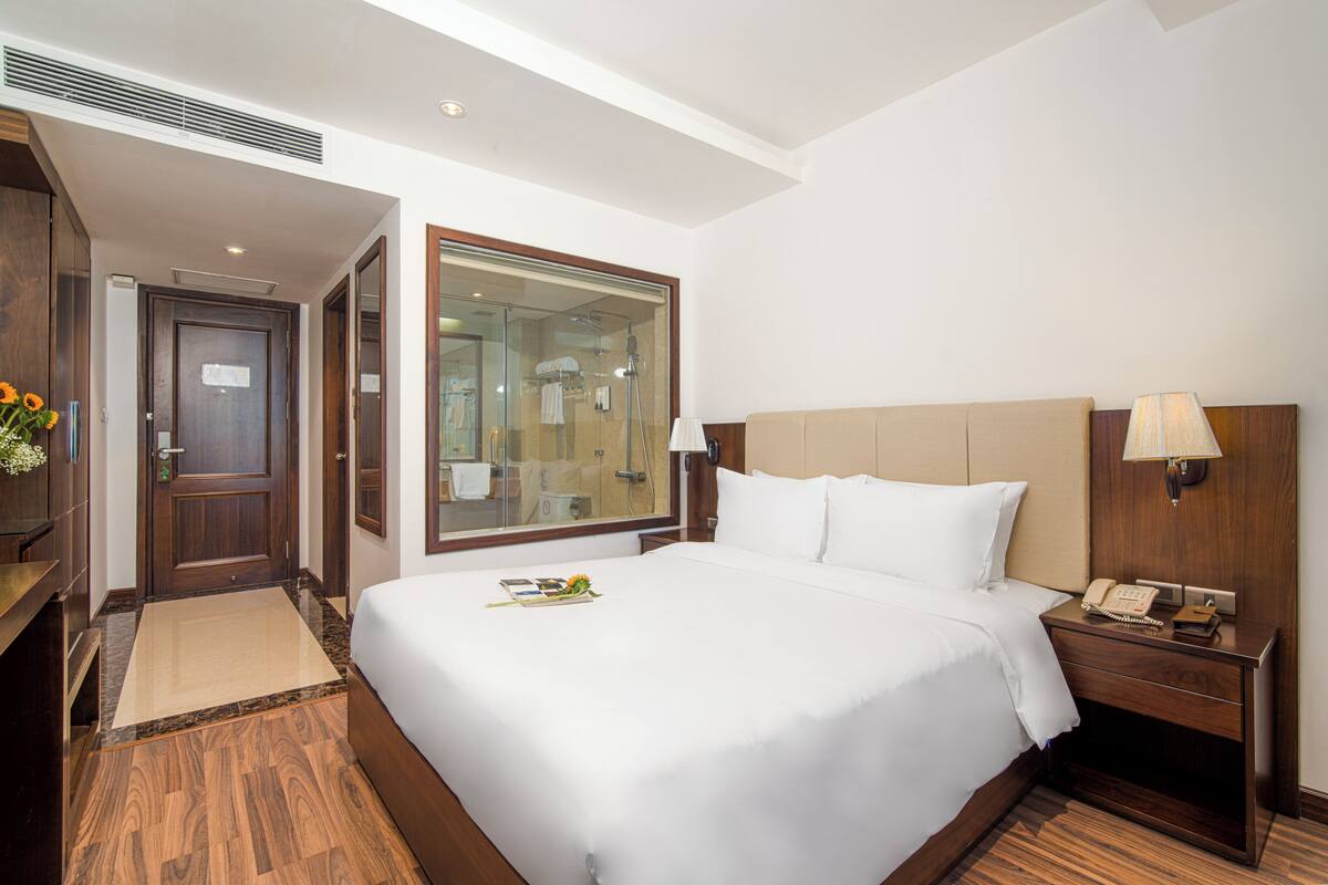 A hotel room with a large bed, a bedside table with a lamp, a wooden nightstand, a wooden wardrobe, a bathroom with a glass shower enclosure, and a small rug.