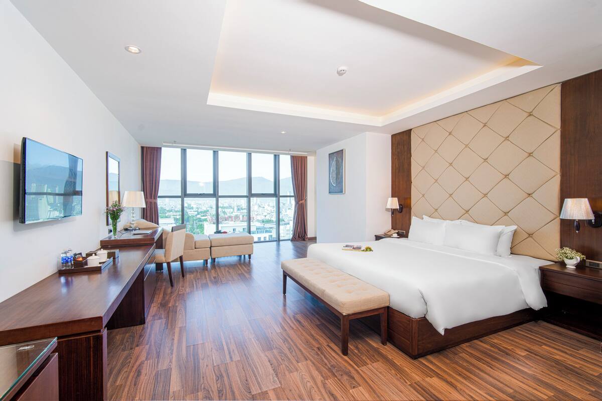 A modern hotel room with a large bed, a desk, a sofa, and a view of the city.