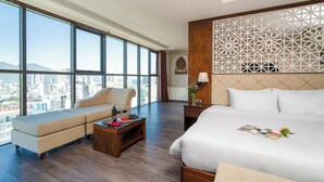 A hotel room with a large bed, a sofa, a wooden coffee table with wine and snacks, and a view of the city through large windows. - Luxtery Hotel (Da Nang)
