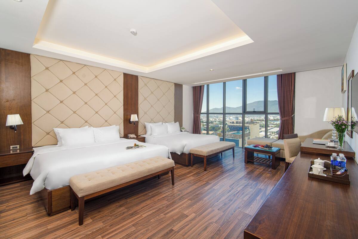 A hotel room with two beds, a sofa, a desk, and a large window offering a city view.