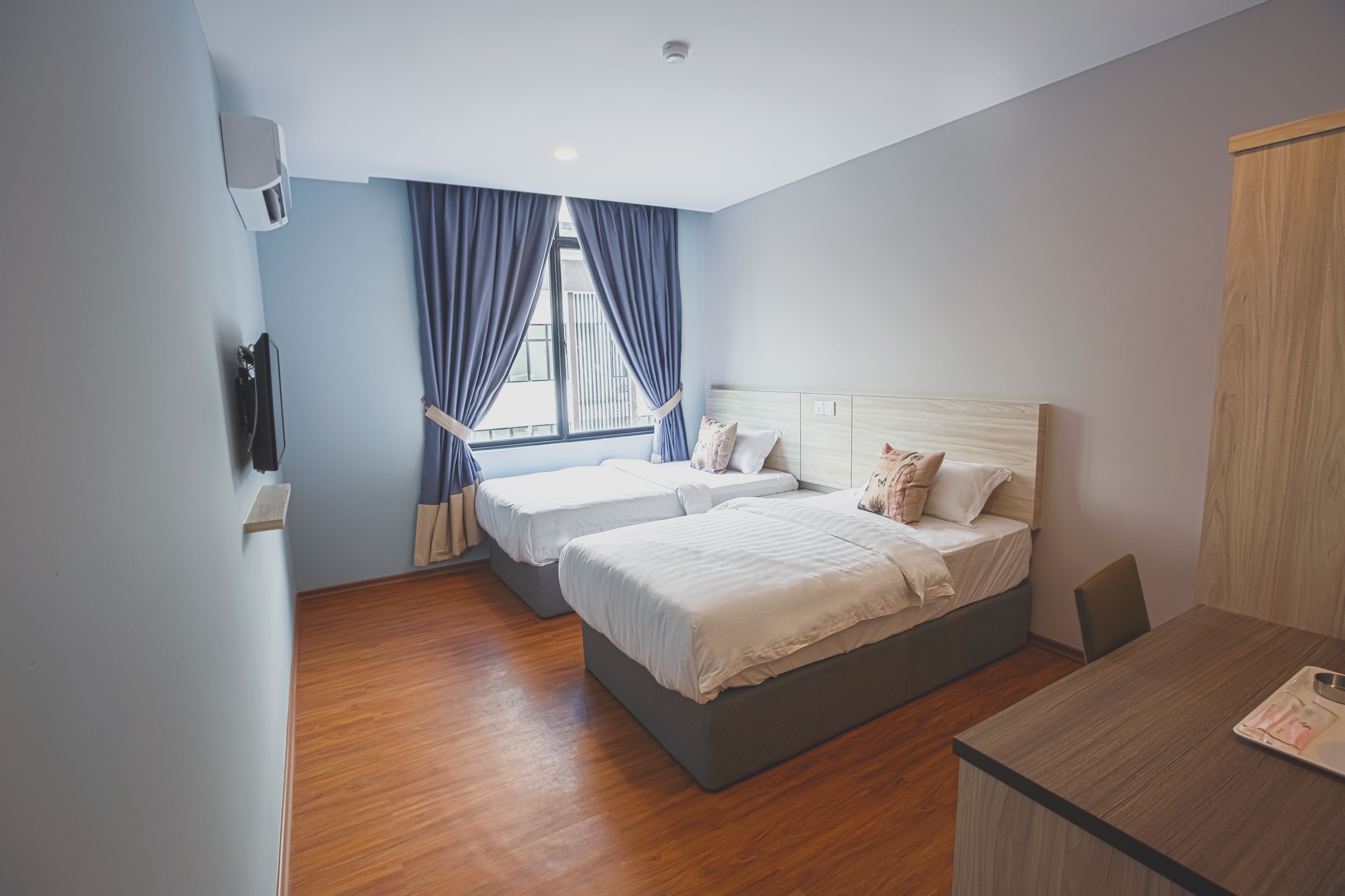 A modern hotel room with two beds, a wooden floor, a desk, and a view of the city through large windows.