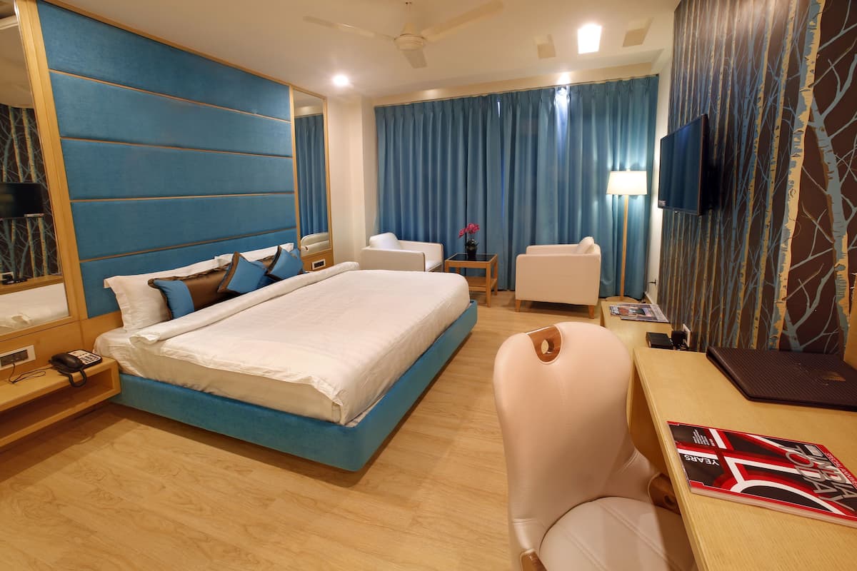 A hotel room with a large bed, a desk with a laptop, a chair, a TV, and a bedside table with a lamp.