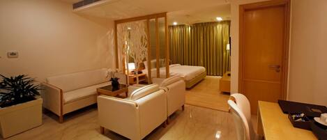 A hotel room with a sofa, armchairs, a dining table, a bed, and a separate sleeping area with curtains.