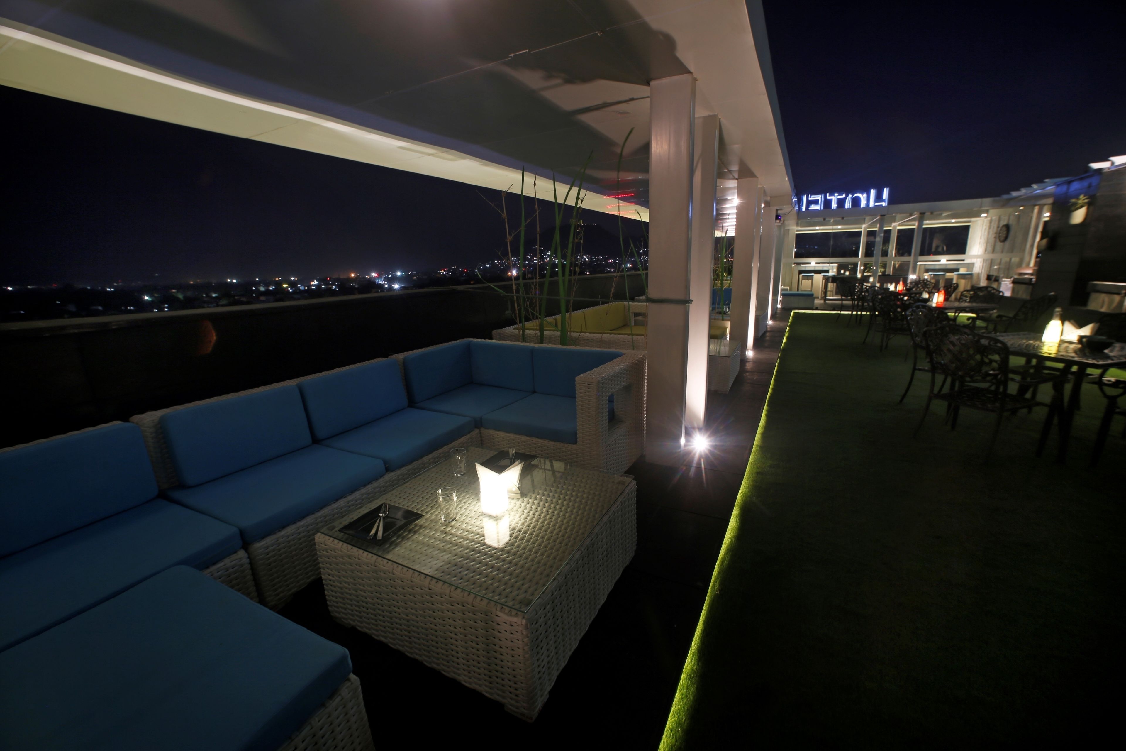 Une terrasse sur le toit avec un canapé bleu, une table en verre dotée d’un éclairage intégré et une vue sur la ville la nuit.