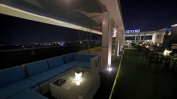 Una terrazza sul tetto con un divano blu, un tavolo con piano in vetro e luce, e vista sulla città di notte.