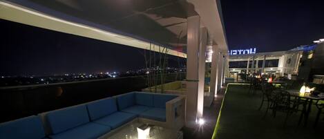 Una terraza en la azotea con un sofá azul, una mesa de vidrio con luz y vistas a la ciudad de noche.