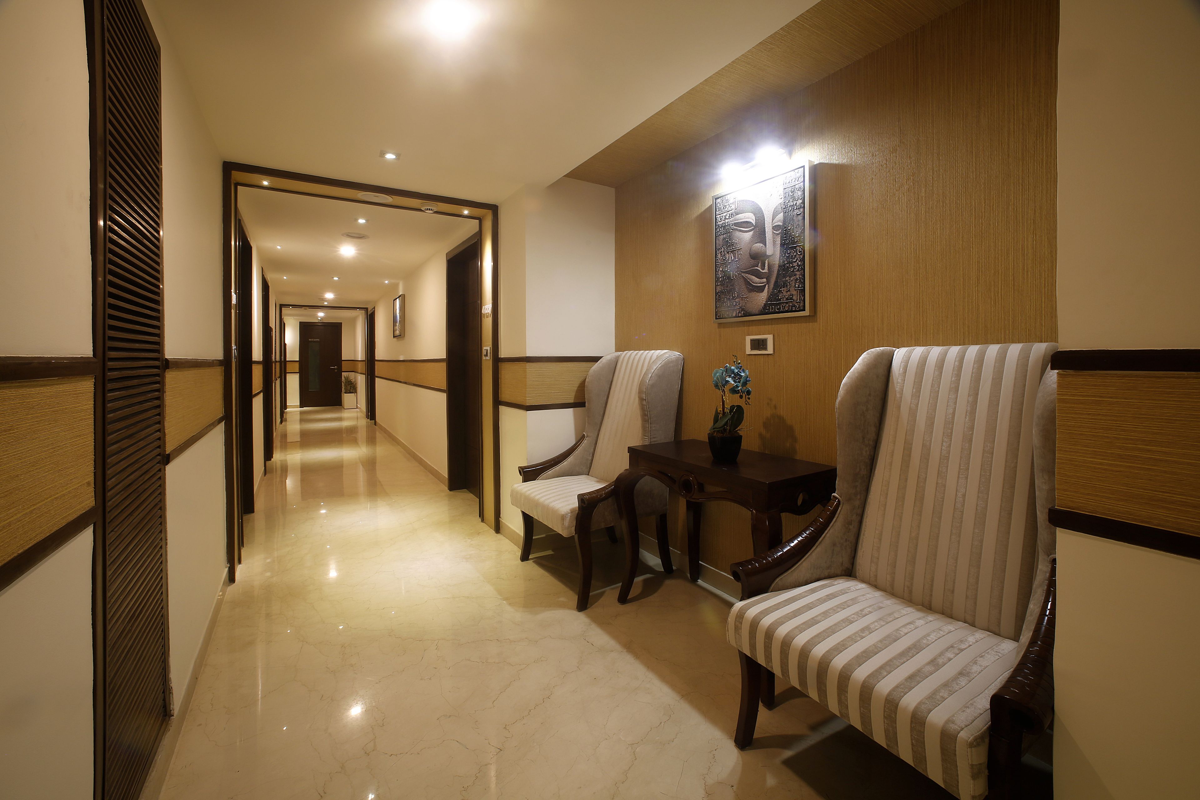 A hotel corridor with a wooden floor, two upholstered chairs, a small table with a potted plant, and a framed artwork on the wall.