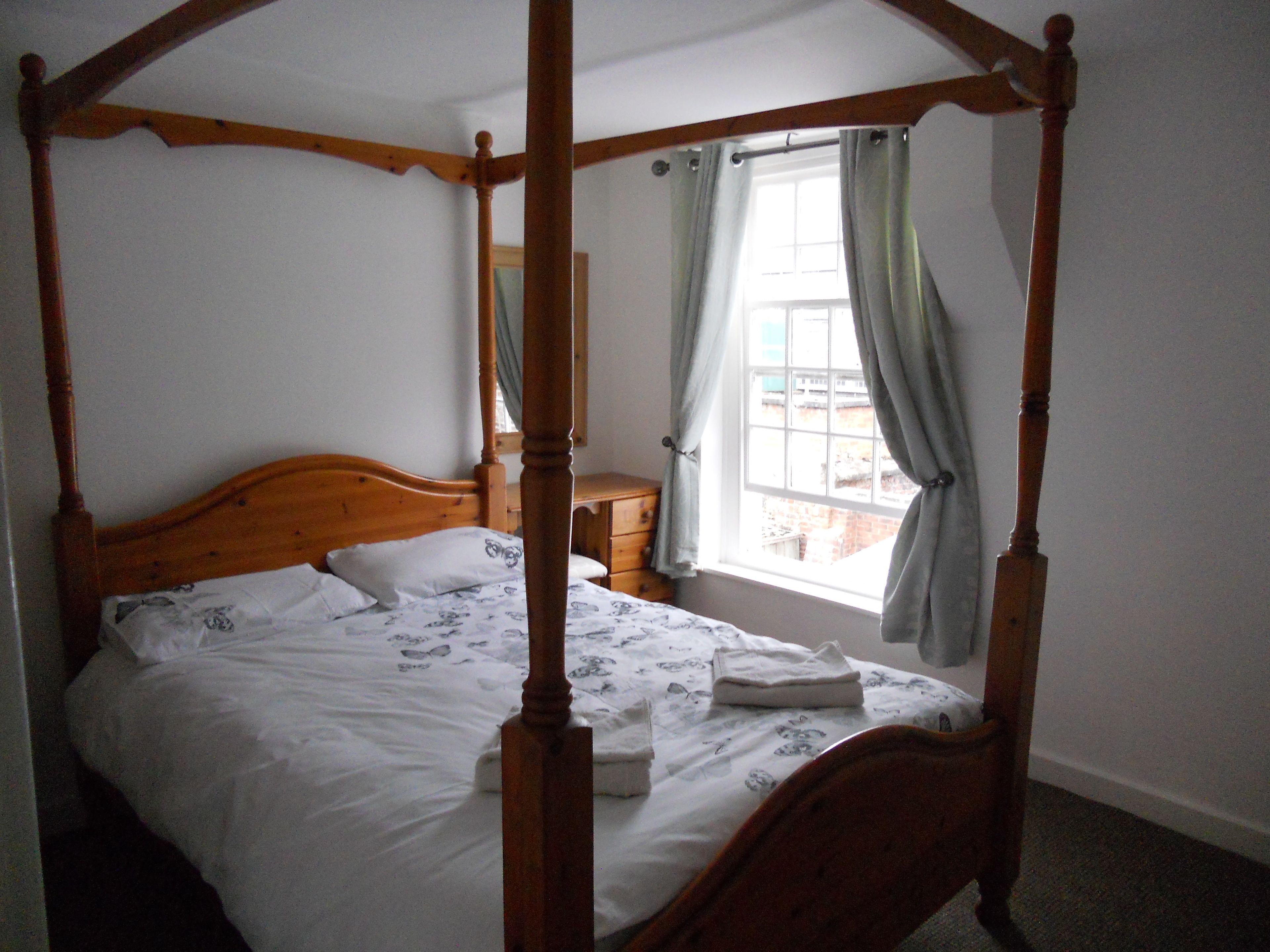 A four-poster bed with a wooden headboard and footboard, a wooden nightstand, and a window with curtains.