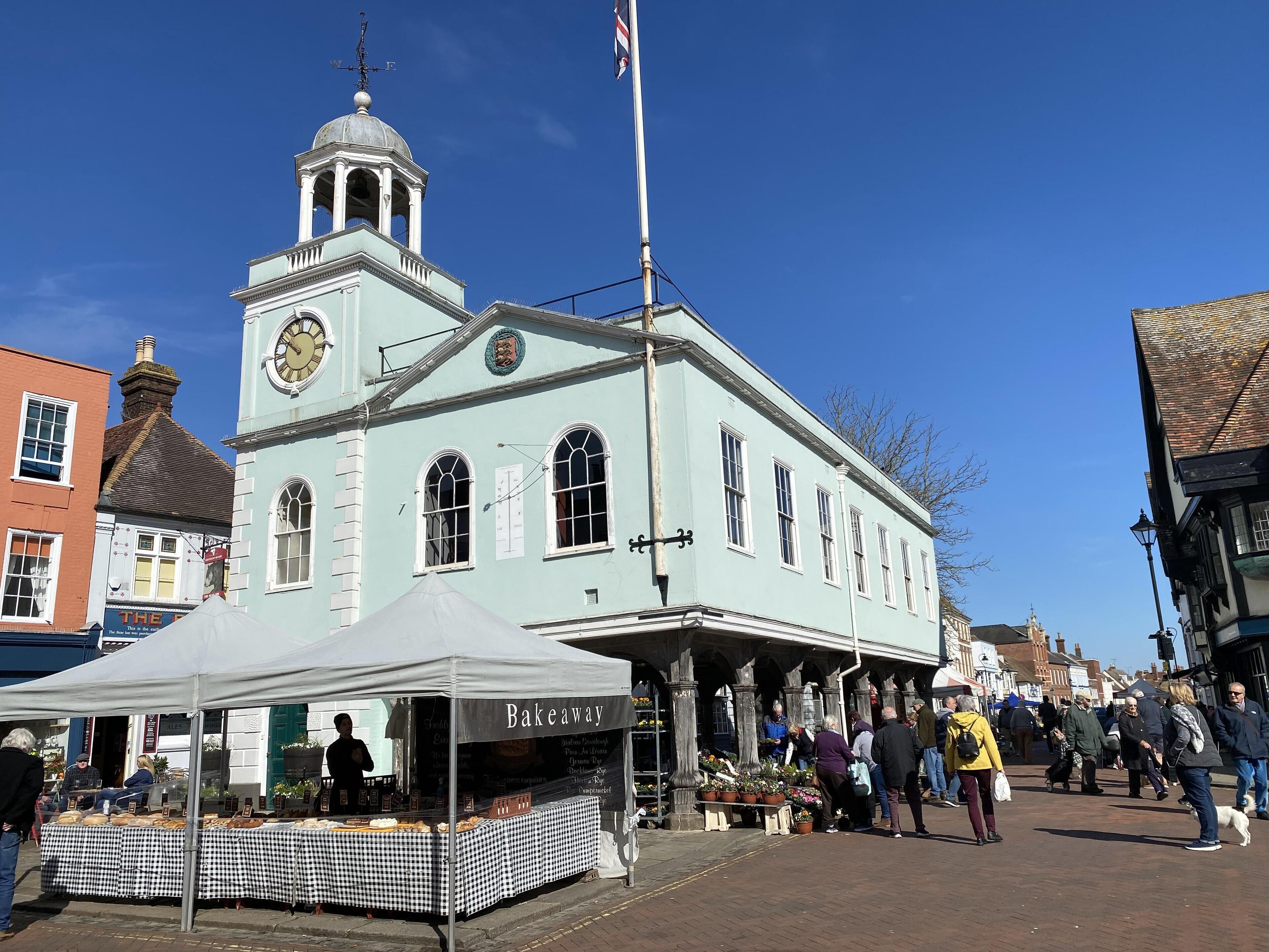A historic building with a clock tower, a market stall, and people walking on the street.