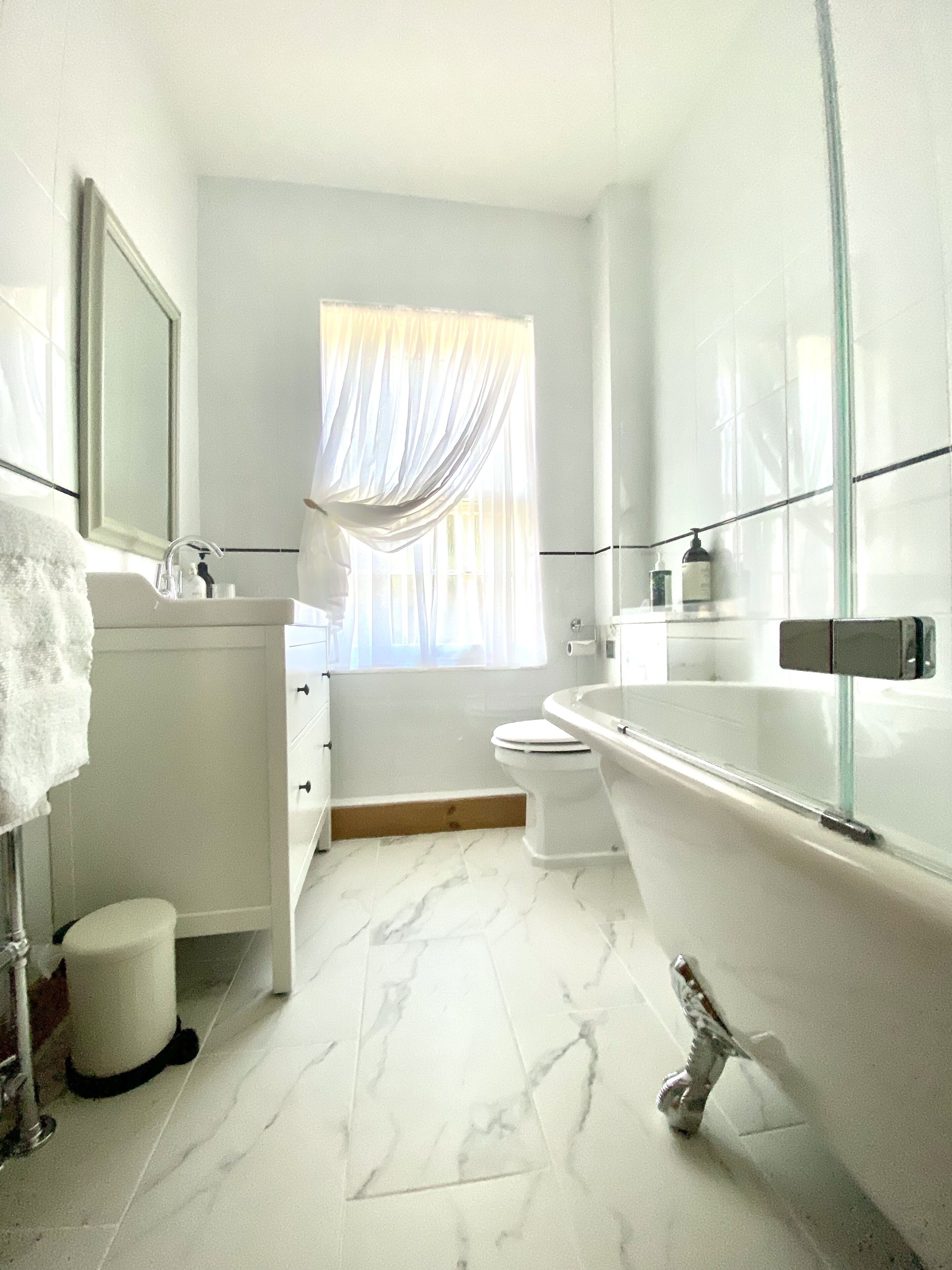 Un baño moderno con tocador blanco, inodoro, bañera y ventana con cortinas traslúcidas.