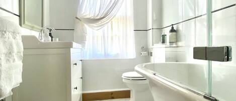 Un baño moderno con tocador blanco, inodoro, bañera y ventana con cortinas traslúcidas.
